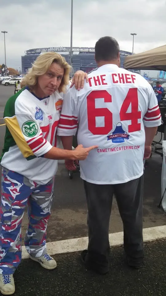 Dan Marney from behind in a white THE CHEF 64 jersey printed with the Gametime Catering logo and web address, while a fan in a Giants jersey points at it, MetLife Stadium behind them.