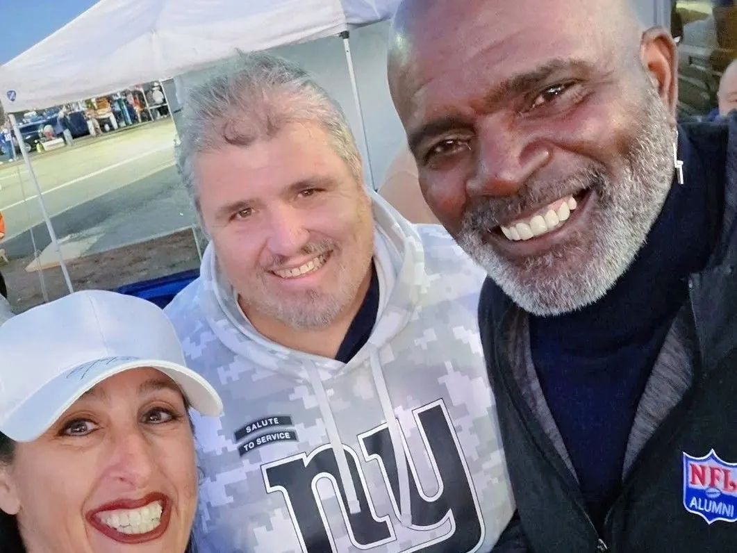 A person in a white cap, Dan Marney in a camouflage-pattern hoodie, and Lawrence Taylor in a dark jacket, close to the camera under a white pop-up canopy with a parking lot behind them