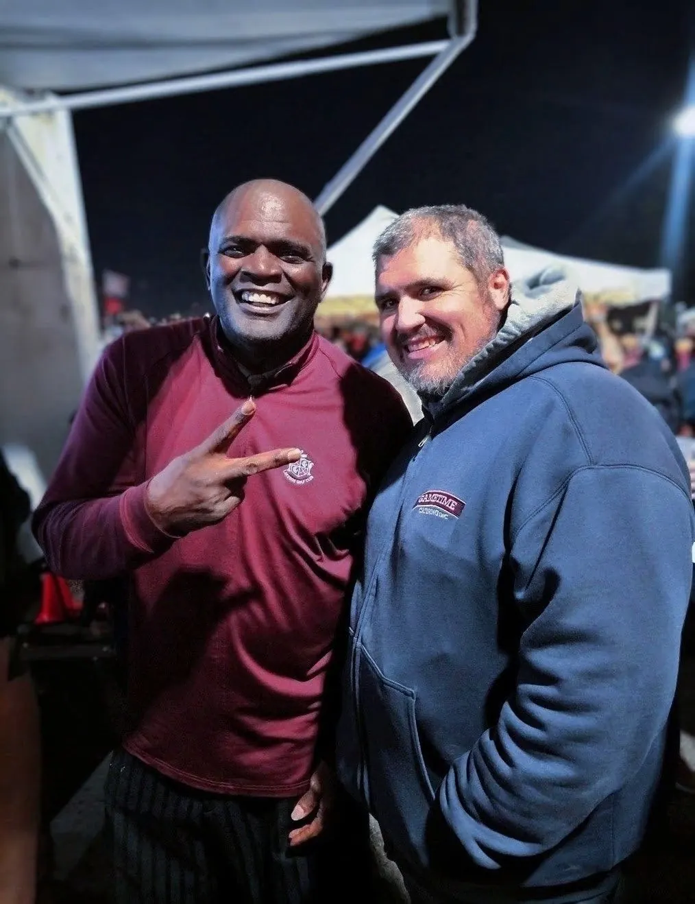 Dan Marney in a blue-grey Gametime Catering hoodie beside Lawrence Taylor, who wears a maroon quarter-zip and holds up two fingers. Both are smiling at the camera under a lit white canopy, with a night crowd behind them.