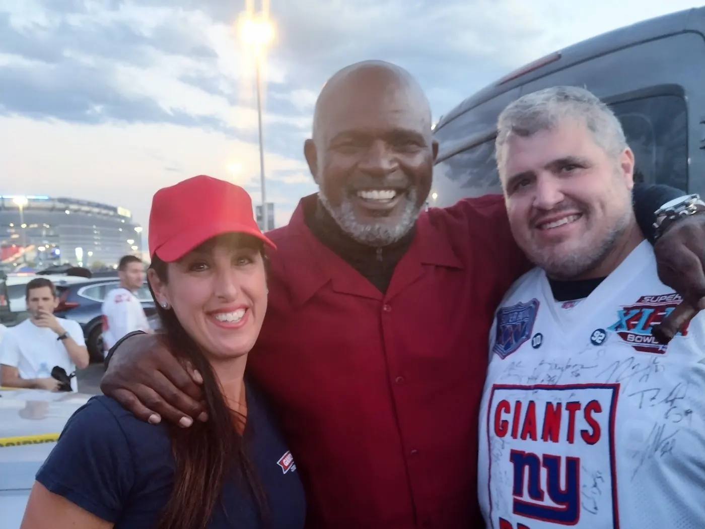 Lawrence Taylor arm in arm with two other people beside a silver-grey coach at golden hour, MetLife Stadium and the lit parking lot behind them.